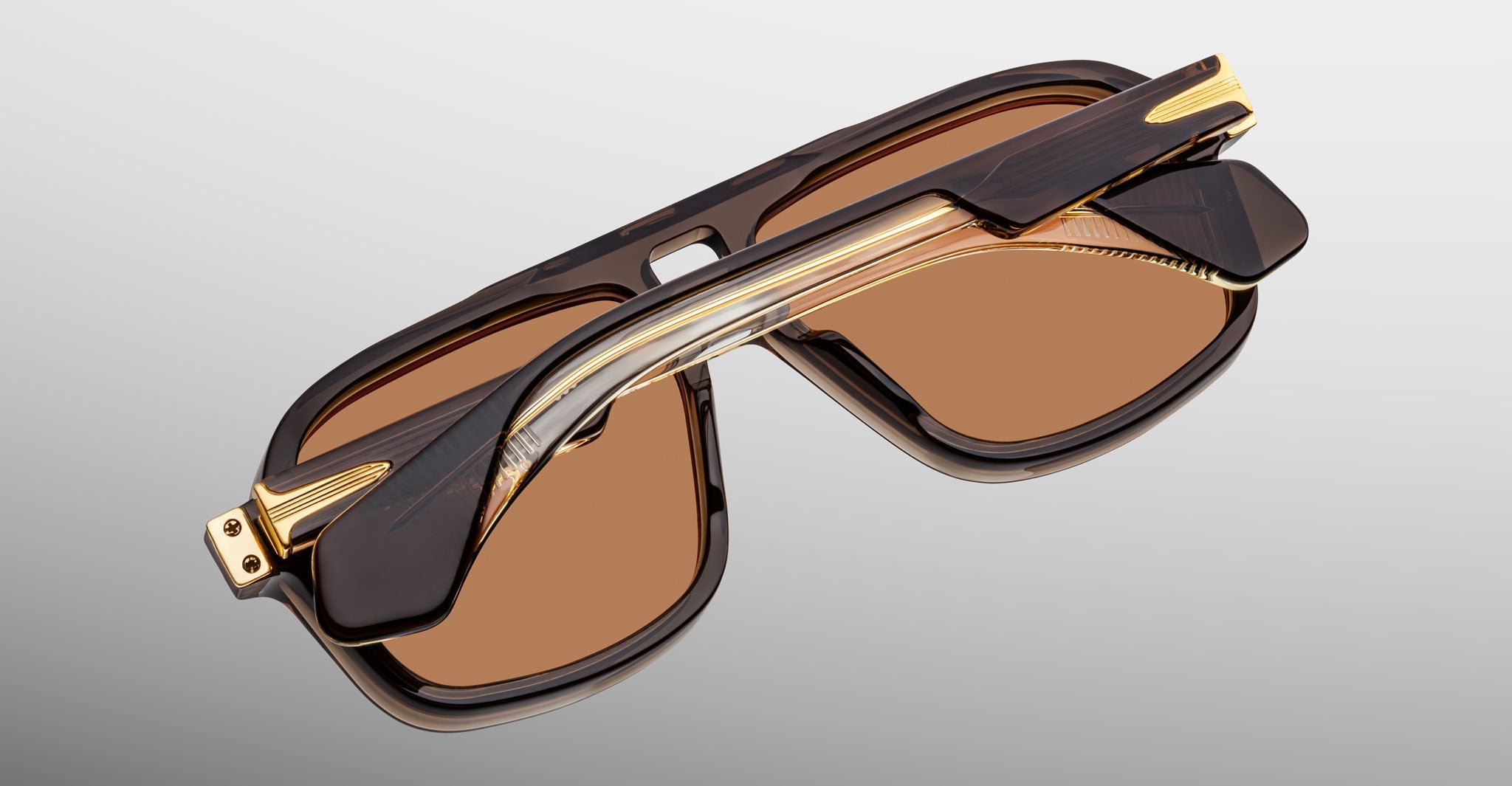 A pair of Jacques Marie Mage Elkhart limited-edition navigator sunglasses with a sculptural brow ridge and amber-tinted lenses, shown from above with crossed arms.