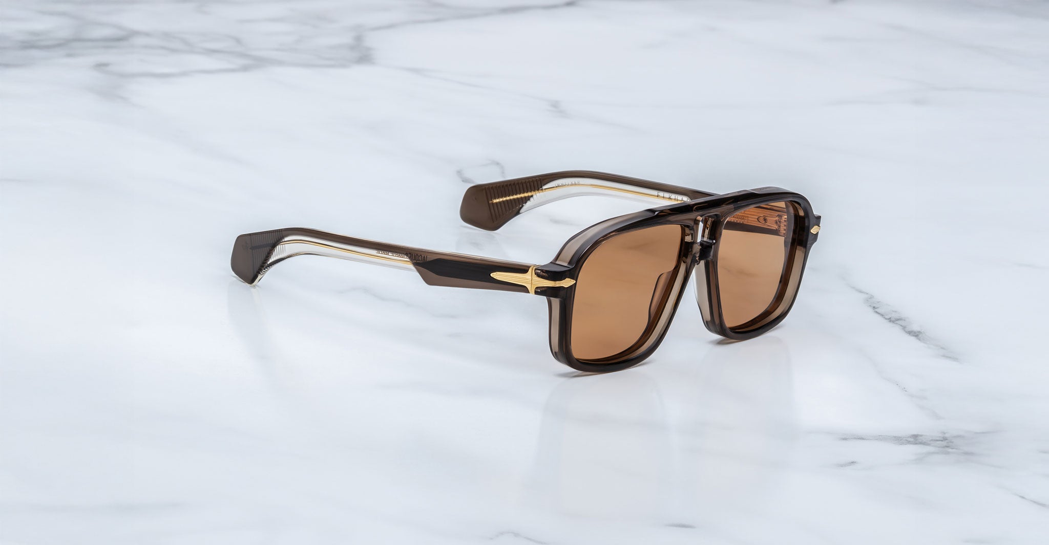A pair of limited-edition Jacques Marie Mage Elkhart navigator sunglasses, with a sculptural brow ridge, thick dark translucent frames, and gold accents, rests on a white marble surface.