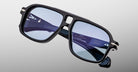 Close-up of Jacques Marie Mage Elkhart navigator sunglasses—black rectangular frames with blue-tinted lenses and a sculptural brow ridge—shown on a light gray background. Limited-edition by Jacques Marie Mage.