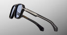Side view of the Jacques Marie Mage Elkhart limited-edition navigator sunglasses, featuring blue-tinted lenses and a sculptural brow ridge, set against a gray gradient background.