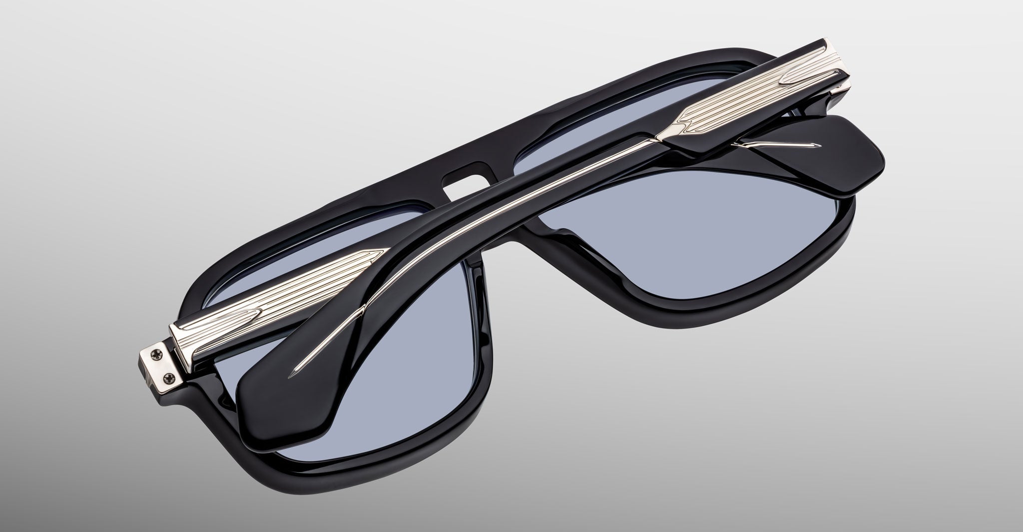 Close-up of Jacques Marie Mage Elkhart sunglasses in black with silver accents and a sculptural brow ridge, folded on a light gray background—a refined, limited-edition navigator from Jacques Marie Mage.