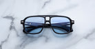 A pair of Jacques Marie Mage Elkhart limited-edition navigator sunglasses with black frames, sculptural brow ridge, and blue-tinted lenses rests on a subtly veined white marble surface.