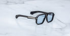 The Jacques Marie Mage Elkhart sunglasses, featuring a black rectangular navigator design with a sculptural brow ridge, blue-tinted lenses, and silver arm accents, are displayed elegantly on a white marble surface.