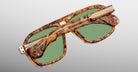 A pair of Jacques Marie Mage Elkhart sunglasses featuring a tortoiseshell pattern, green lenses, and a sculptural brow ridge, folded on a plain light gray background.