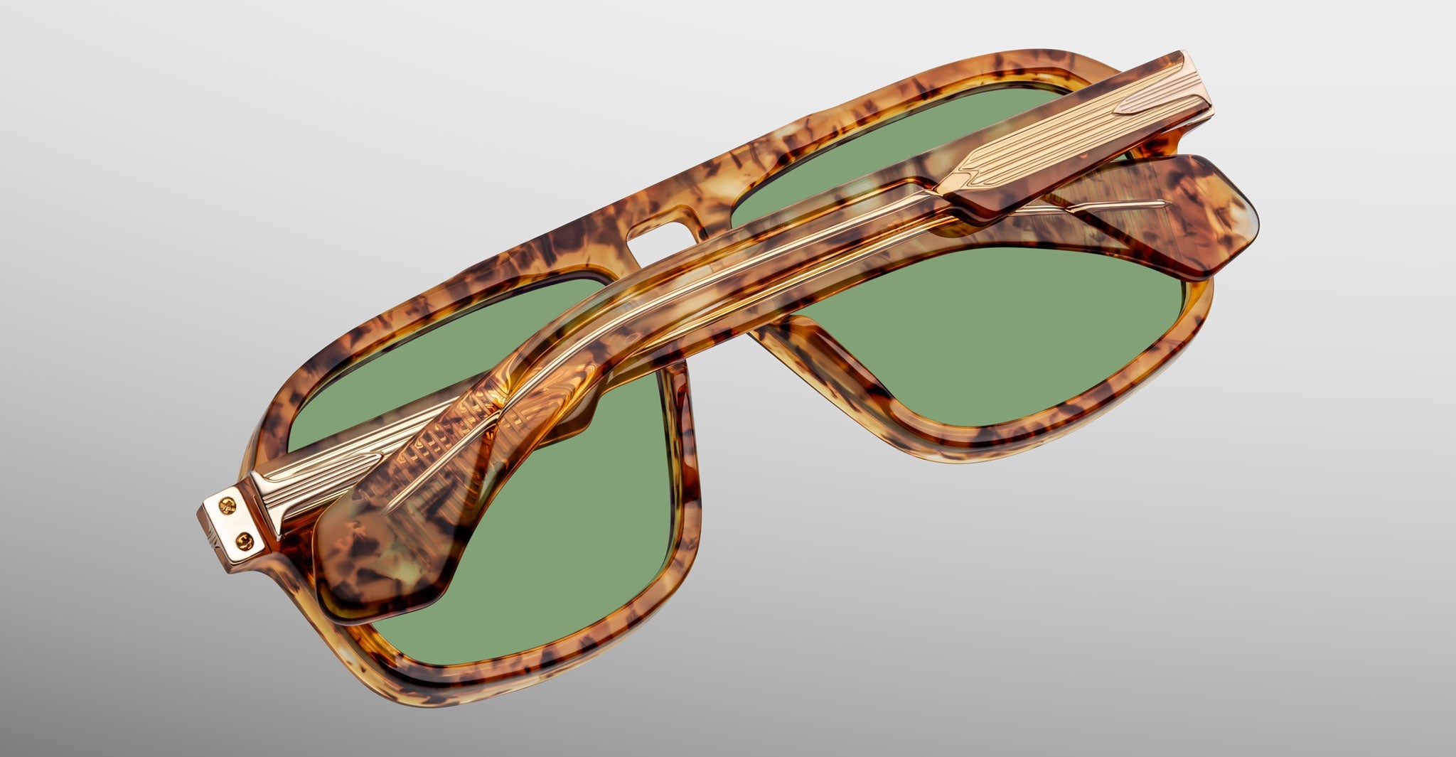 A pair of Jacques Marie Mage Elkhart sunglasses featuring a tortoiseshell pattern, green lenses, and a sculptural brow ridge, folded on a plain light gray background.