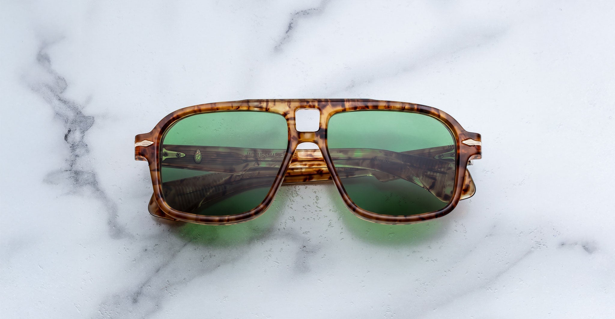 The Jacques Marie Mage Elkhart tortoiseshell sunglasses, featuring green lenses and a sculptural brow ridge, are displayed on a white marble surface.