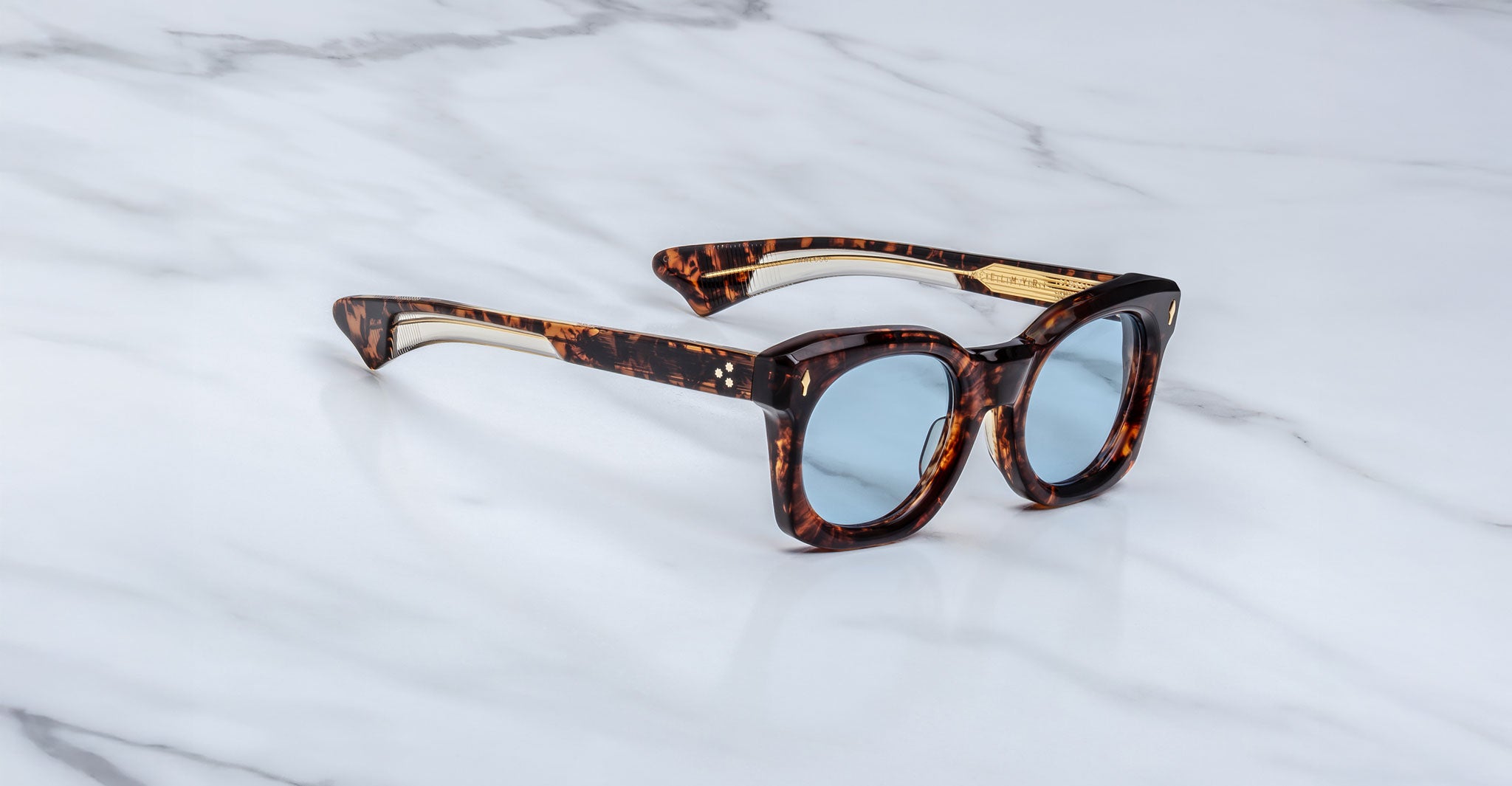 The Jacques Marie Mage Small Elmyr sunglasses, featuring thick tortoiseshell frames and blue-tinted lenses, rest on a white marble surface.