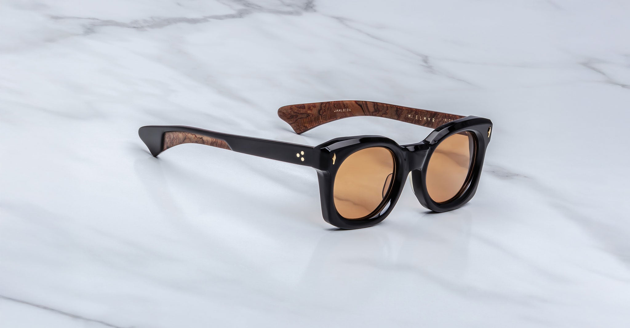 The Jacques Marie Mage Small Elmyr sunglasses feature round brown lenses, thick black frames, and brown wood-textured inner arms, shown here resting on a white marble surface.