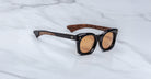 The Jacques Marie Mage Small Elmyr sunglasses feature round brown lenses, thick black frames, and brown wood-textured inner arms, shown here resting on a white marble surface.