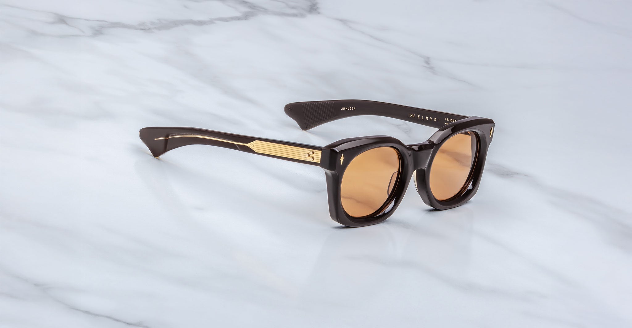 The Jacques Marie Mage Small Elmyr sunglasses feature dark brown frames with gold-accented arms and round brown-tinted lenses, displayed on a white marble surface.