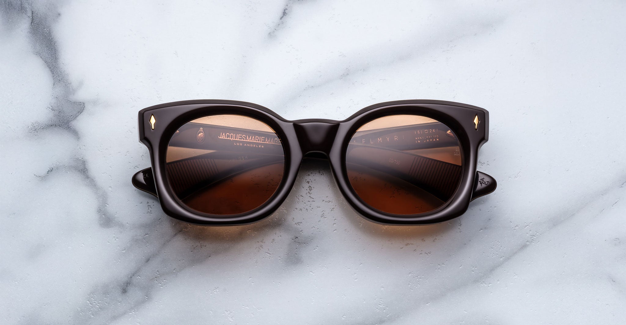 A pair of Jacques Marie Mage Small Elmyr sunglasses, featuring dark brown thick frames and brown-tinted lenses, rests on a white marble surface. Some text is reflected on the inside of the left lens.
