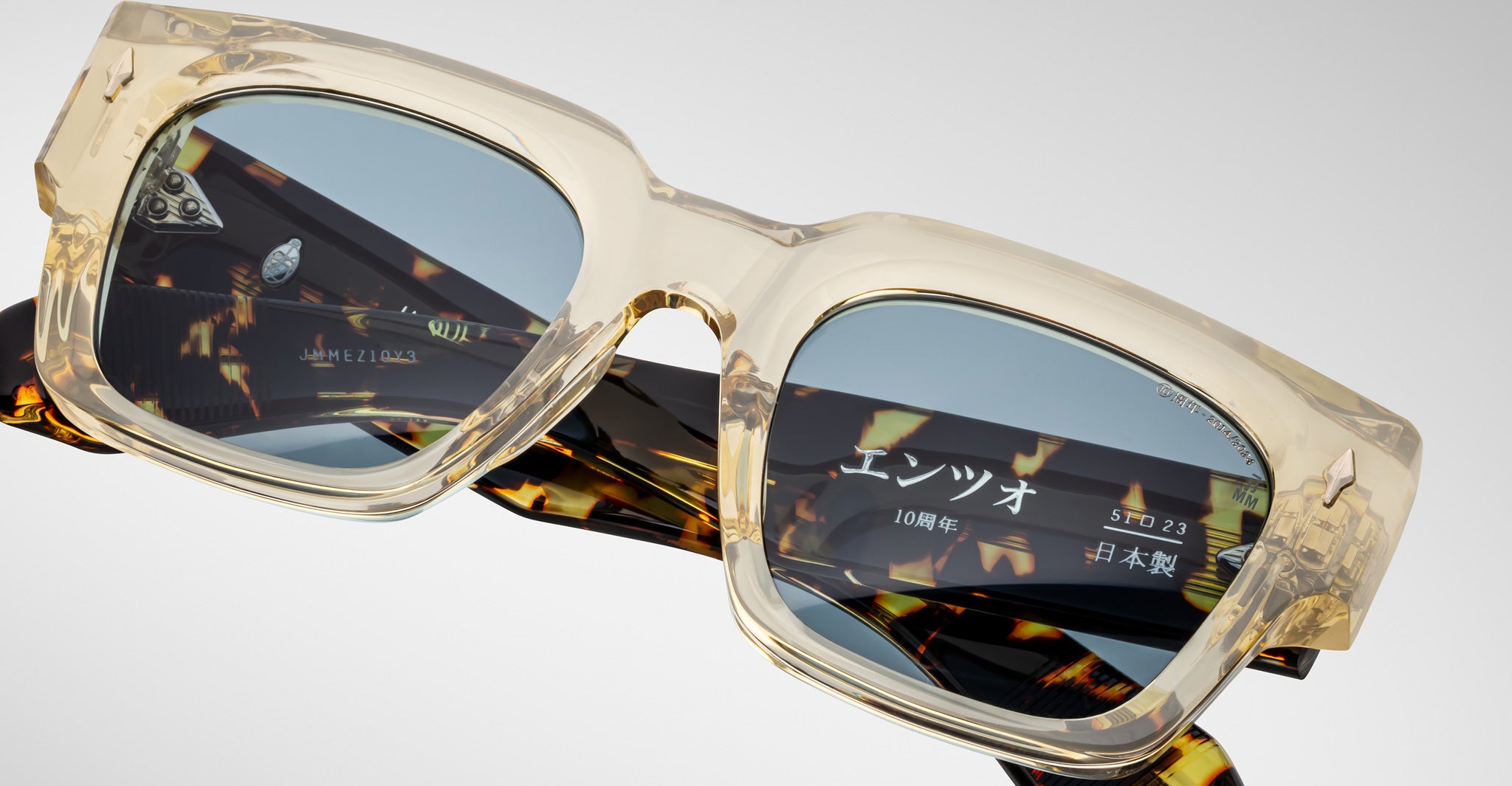 Close-up of Jacques Marie Mage Enzo sunglasses with clear frames, blue lenses, and tortoiseshell arms, viewed from the side.