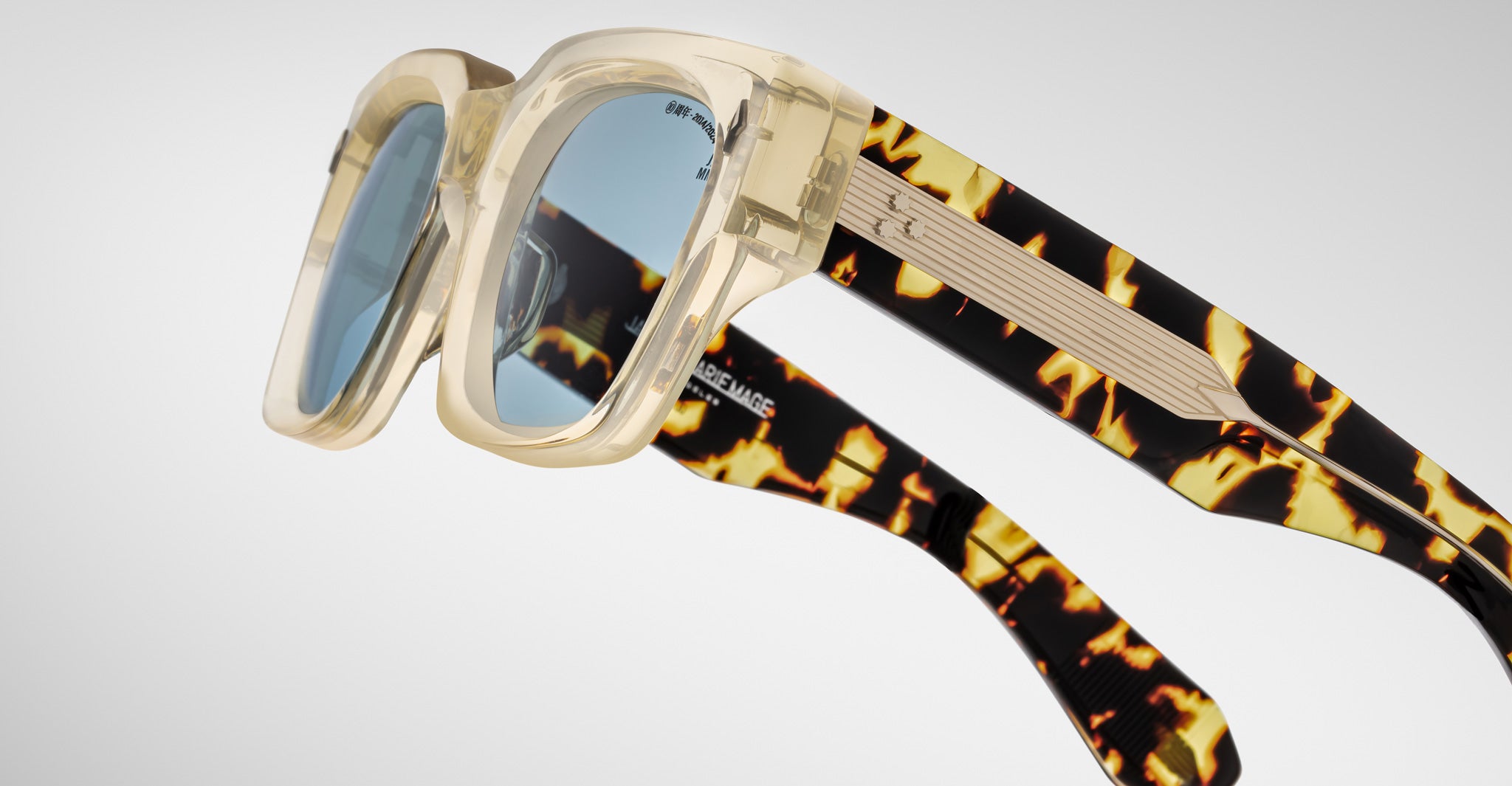 Jacques Marie Mage Enzo sunglasses with clear frame, blue lenses, and tortoiseshell arms shown from a front view.