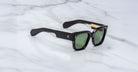 Jacques Marie Mage Enzo black rectangular sunglasses with green lenses and gold accents, shown from above.