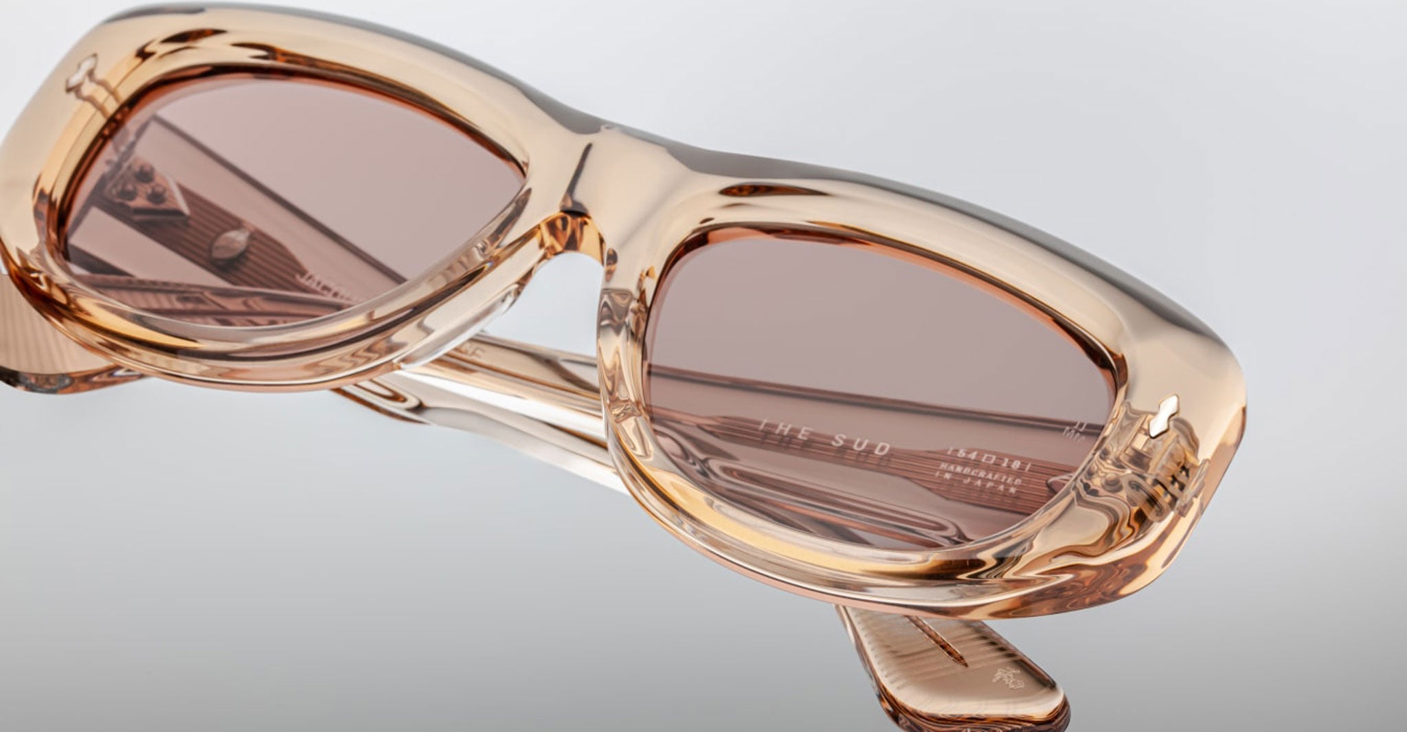 Close-up of Jacques Marie Mage Erin Wasson - Sud sunglasses with beige-tinted frames and brown lenses, resting upside down on a reflective surface, highlighting the detailed texture and subtle inner arm branding.