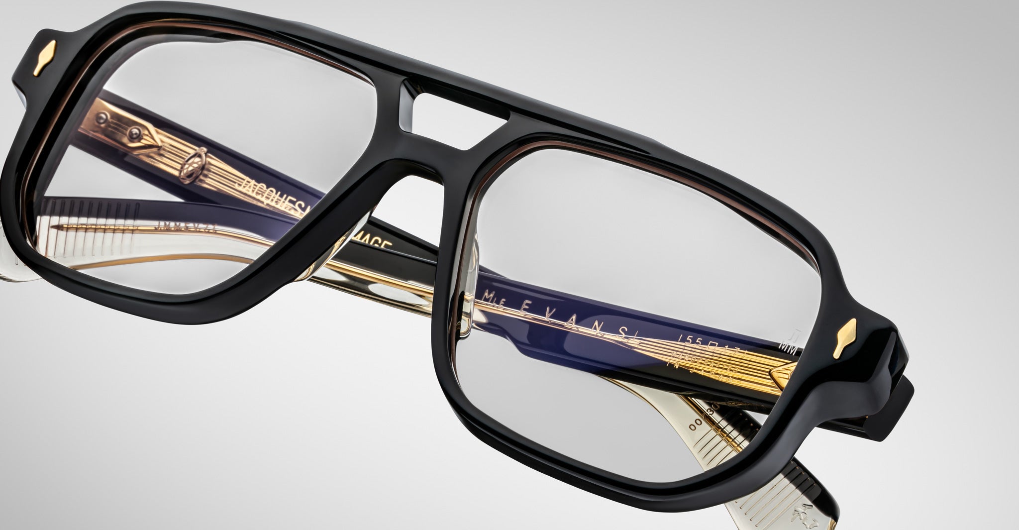 Close-up of Jacques Marie Mage Evans handcrafted optical glasses with an oversized acetate frame and gold and dark blue temple accents, set against a plain light background.