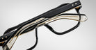 Two pairs of modern eyeglasses, including the Jacques Marie Mage Evans oversized acetate frame in black and gold, are stacked together on a plain gray background.
