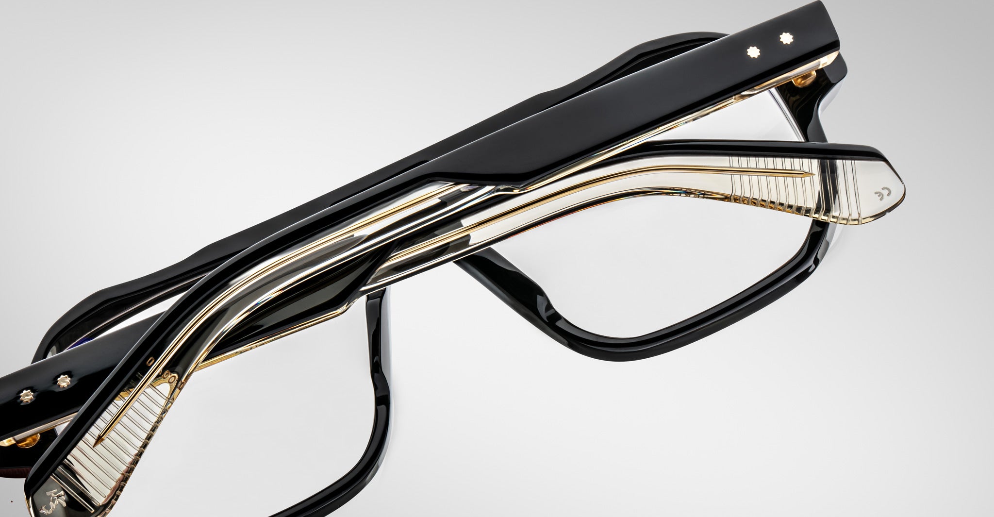 Two pairs of modern eyeglasses, including the Jacques Marie Mage Evans oversized acetate frame in black and gold, are stacked together on a plain gray background.
