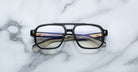 Jacques Marie Mage Evans: Oversized black-rimmed handcrafted optical glasses with gold and navy blue acetate arms by Jacques Marie Mage, displayed on a white marble surface.