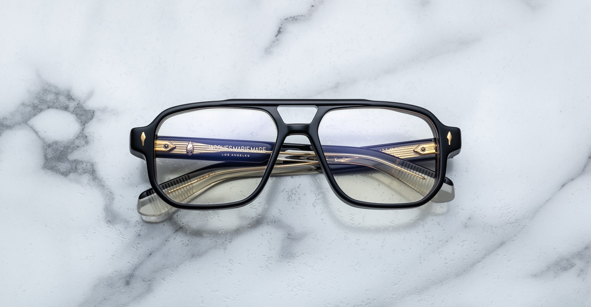 Jacques Marie Mage Evans: Oversized black-rimmed handcrafted optical glasses with gold and navy blue acetate arms by Jacques Marie Mage, displayed on a white marble surface.