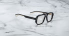 The Jacques Marie Mage Evans optical glasses feature an oversized black and gold acetate frame with clear lenses, showcased on a white marble surface.