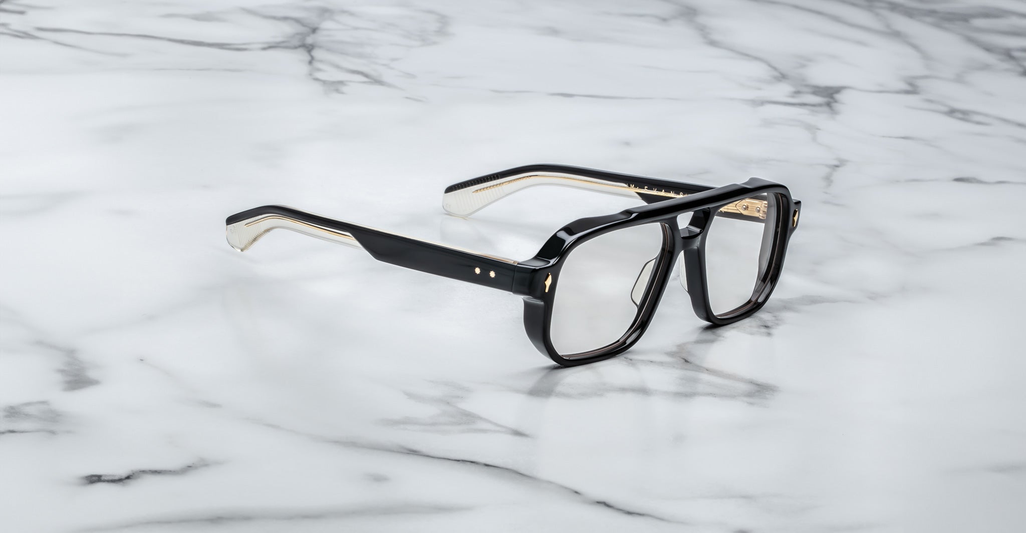 The Jacques Marie Mage Evans optical glasses feature an oversized black and gold acetate frame with clear lenses, showcased on a white marble surface.