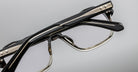 Close-up of Jacques Marie Mage Evans optical glasses, showcasing a black and gold oversized acetate frame with geometric lines and clear lenses, set against a plain gray background.