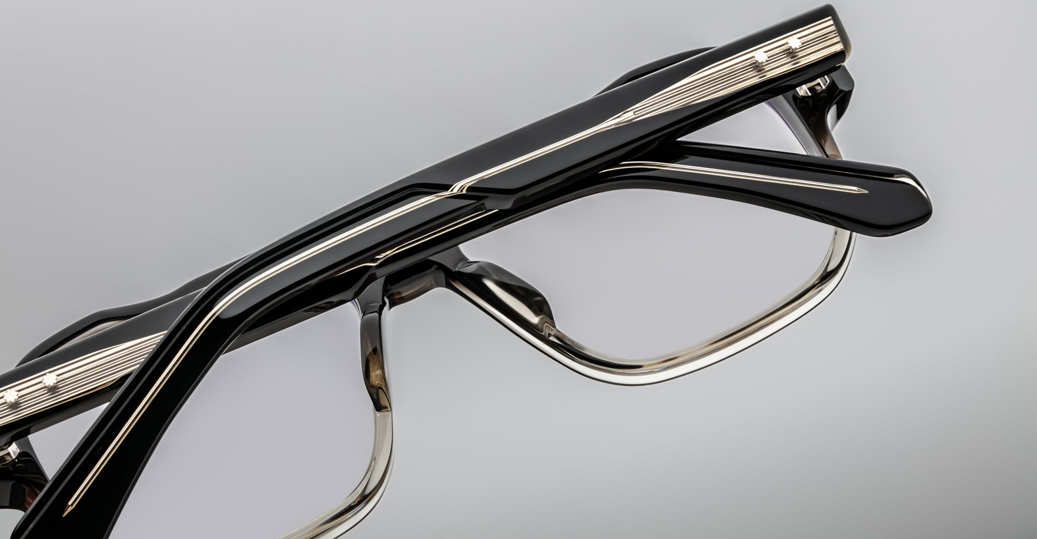 Close-up of Jacques Marie Mage Evans optical glasses, showcasing a black and gold oversized acetate frame with geometric lines and clear lenses, set against a plain gray background.