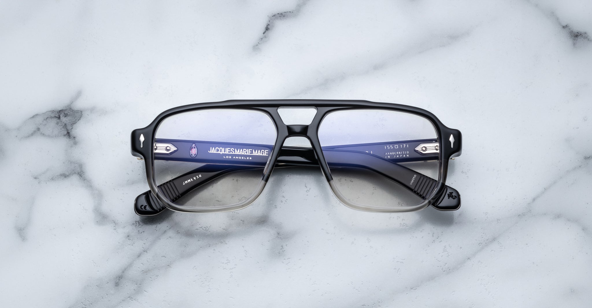 A pair of Jacques Marie Mage Evans eyeglasses rests on white marble, featuring oversized handcrafted acetate frames with the Jacques Marie Mage branding visible inside.