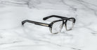 The Jacques Marie Mage Evans eyeglasses by Jacques Marie Mage feature a black and clear ombré oversized acetate frame with thick rectangular silhouette and silver temple detailing, handcrafted and displayed on a white marble surface.