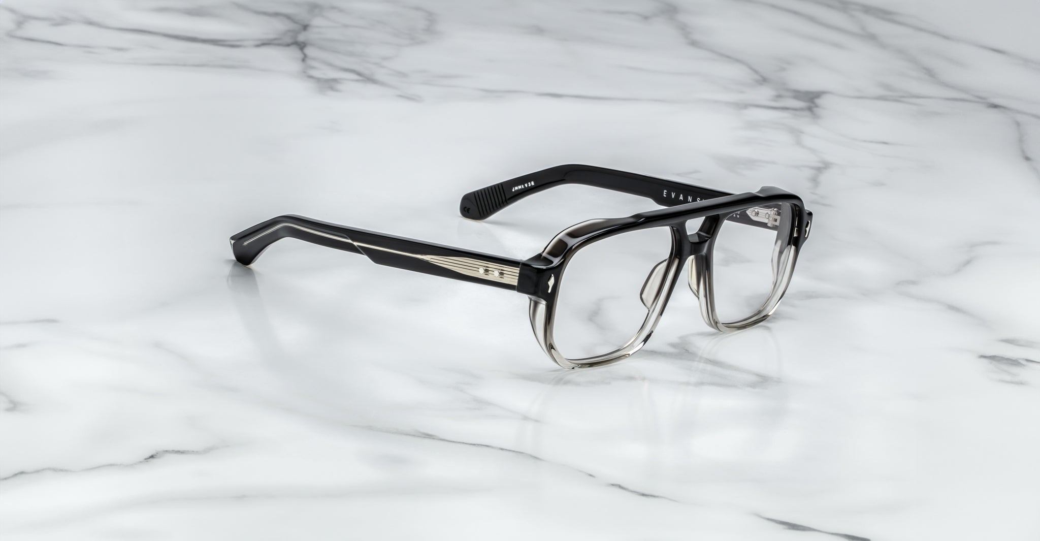 The Jacques Marie Mage Evans eyeglasses by Jacques Marie Mage feature a black and clear ombré oversized acetate frame with thick rectangular silhouette and silver temple detailing, handcrafted and displayed on a white marble surface.