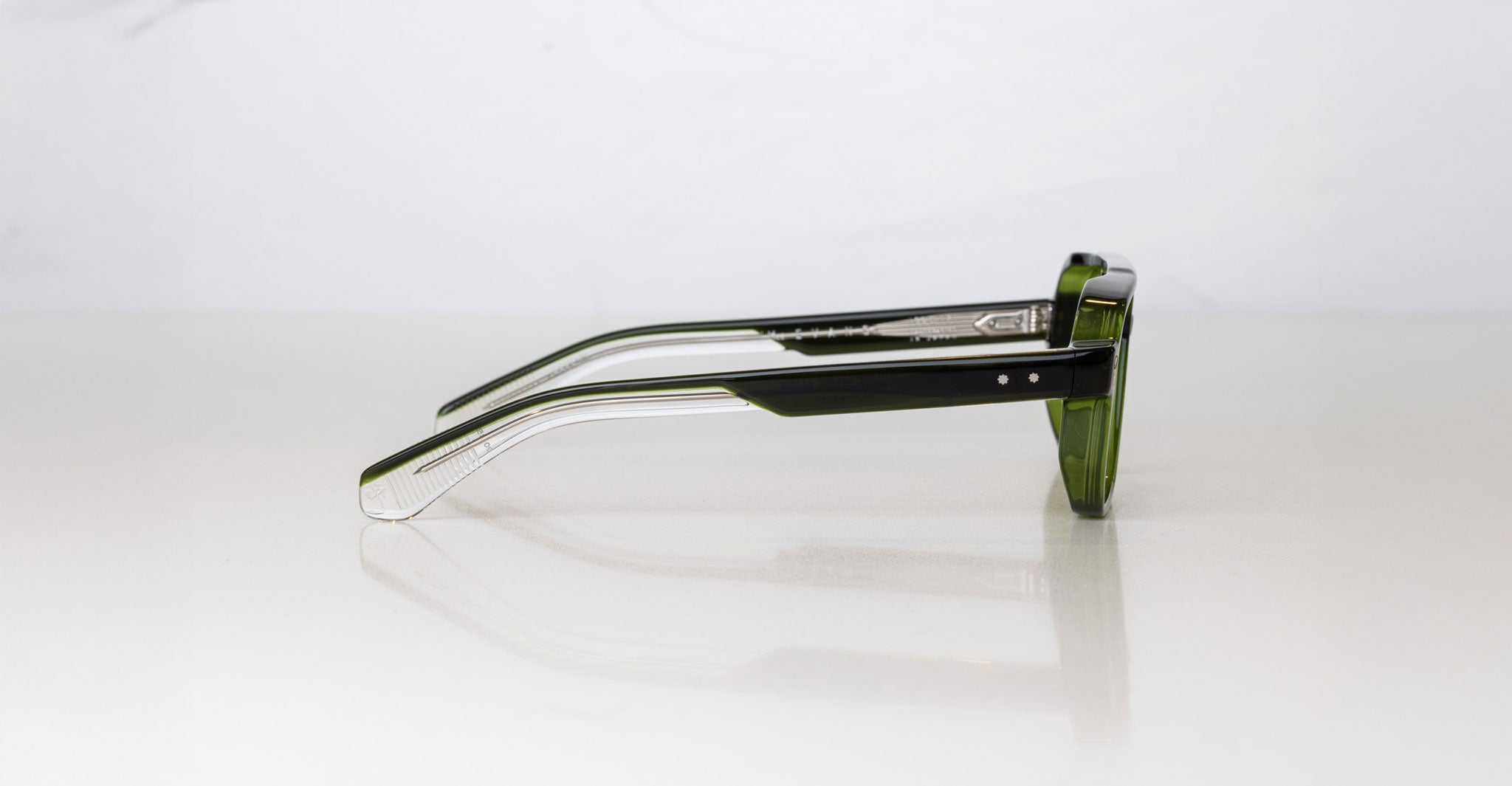 A pair of Jacques Marie Mage Evans green-framed handcrafted optical glasses rests on its side against a plain white background.