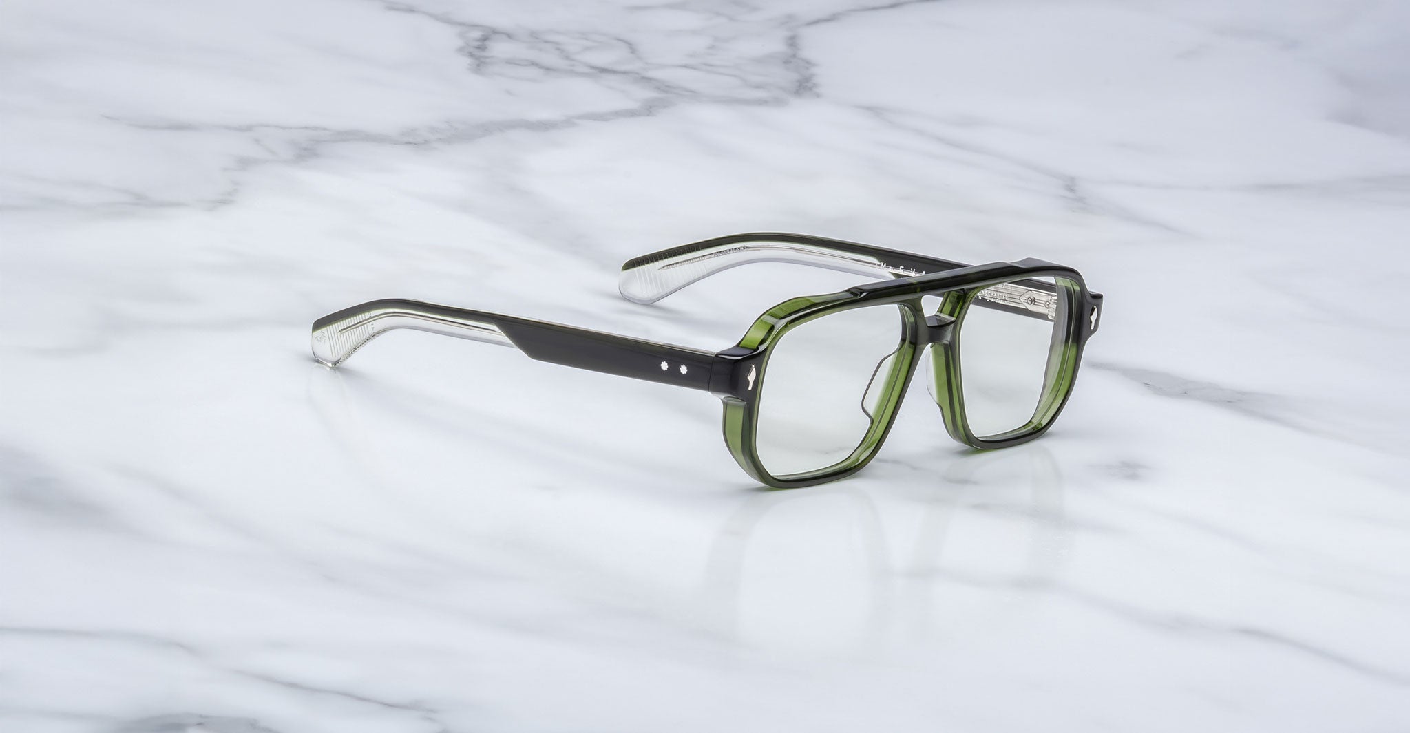 A pair of Jacques Marie Mage Evans handcrafted optical glasses in green and black with thick frames rests on a white marble surface.