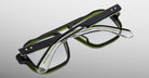 The Jacques Marie Mage Evans optical glasses feature handcrafted thick black and green frames, shown folded with crossed arms against a neutral gradient background.