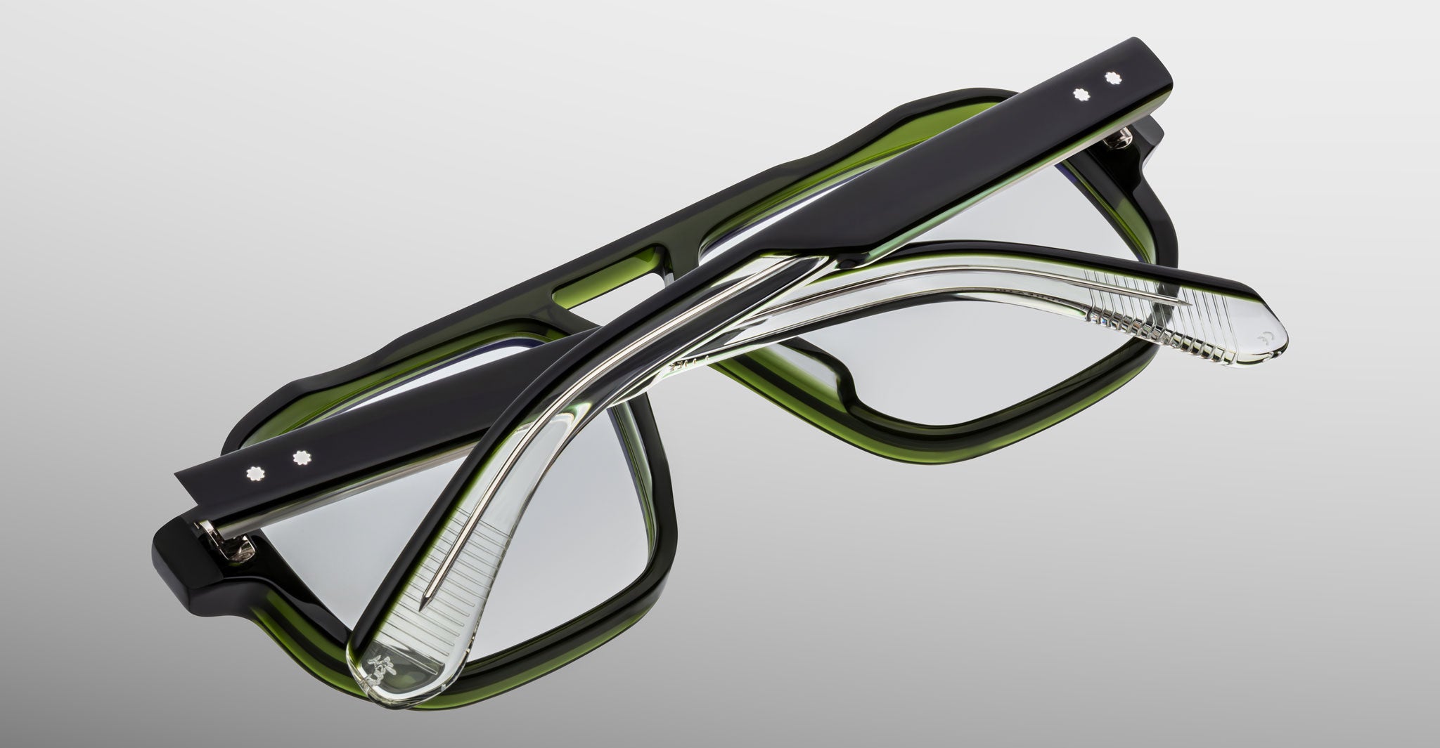 The Jacques Marie Mage Evans optical glasses feature handcrafted thick black and green frames, shown folded with crossed arms against a neutral gradient background.
