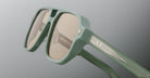 Close-up of Jacques Marie Mage Evans sunglasses in mint green with an oversized acetate frame, gold accents, and light brown lenses set against a gradient gray background.
