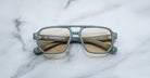 The Jacques Marie Mage Evans eyeglasses by Jacques Marie Mage feature an oversized matte green acetate rectangular frame with gold accents, displayed on a white marble surface.