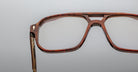 Close-up of handcrafted Jacques Marie Mage Evans optical glasses featuring large, brown, wood-patterned frames, set against a plain light gray background.