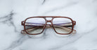 Jacques Marie Mage Evans handcrafted optical glasses with oversized brown acetate frames and clear lenses rest on a white marble surface, their folded arms neatly placed behind the frames.