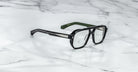 The Jacques Marie Mage Evans glasses feature handcrafted rectangular lenses, silver temple accents, and green inner arms, showcased on a white marble surface.