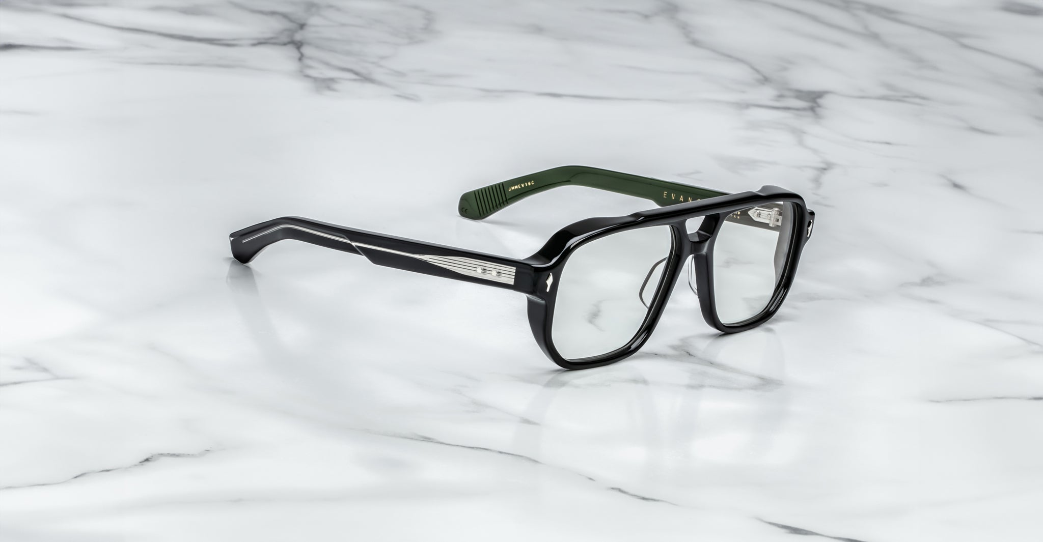 The Jacques Marie Mage Evans glasses feature handcrafted rectangular lenses, silver temple accents, and green inner arms, showcased on a white marble surface.