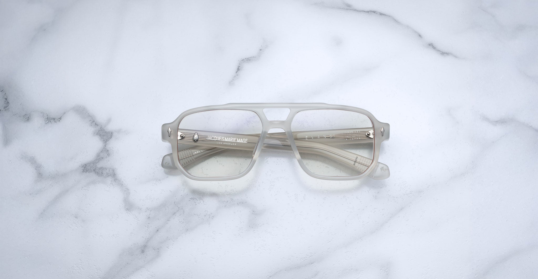 The Jacques Marie Mage Evans eyeglasses by Jacques Marie Mage feature oversized, translucent square acetate frames and are displayed on a white marble surface.