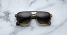 A pair of Jacques Marie Mage Evans sunglasses with dark lenses, an oversized acetate frame, and a greenish tint rests on a white marble surface.