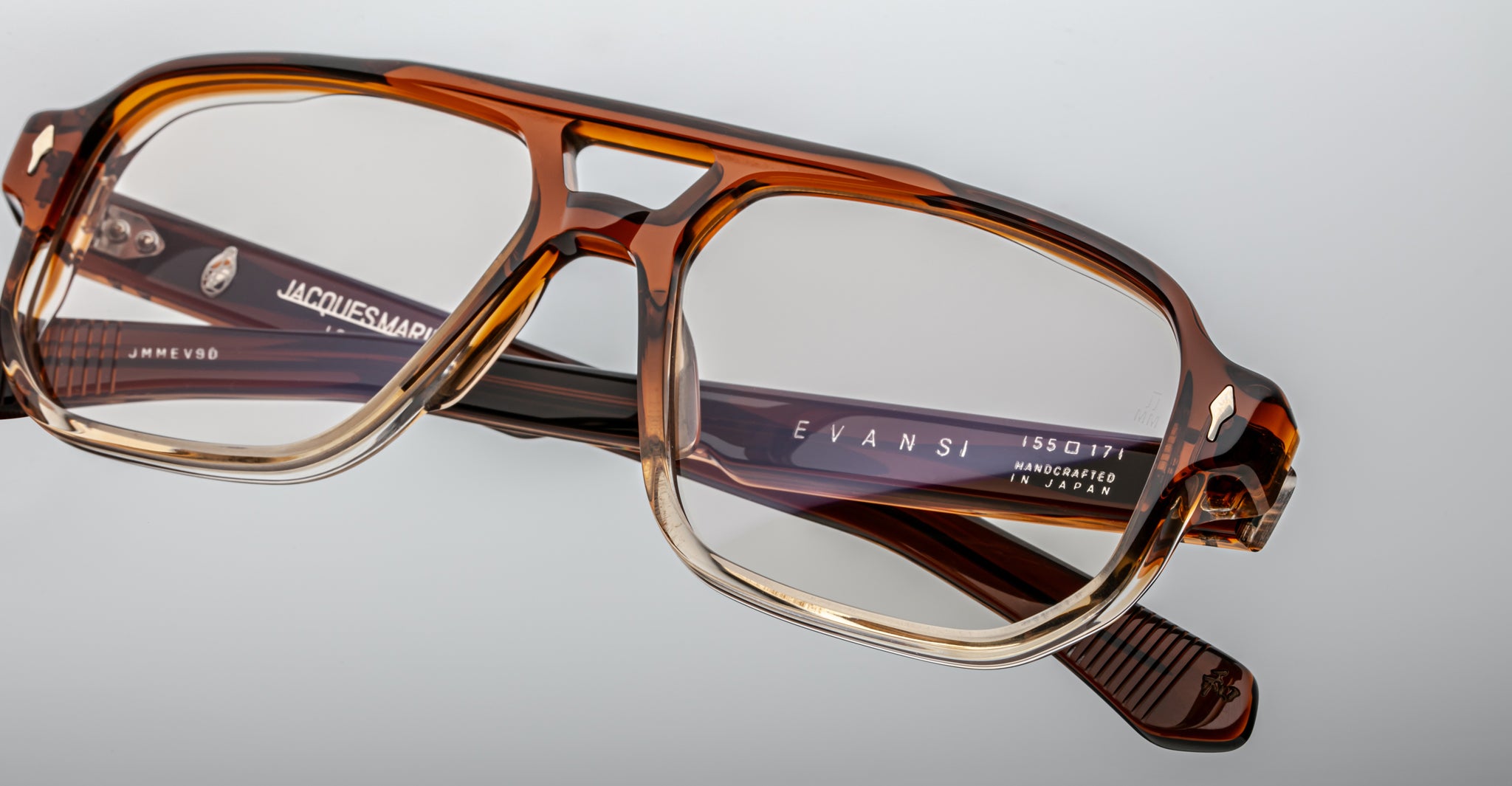 Close-up of handcrafted Jacques Marie Mage Evans optical glasses with a brown and clear oversized acetate frame, featuring the Jacques Marie Mage logo on the temples, set against a plain background.