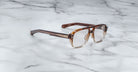 A pair of Jacques Marie Mage Evans eyeglasses with brown, square acetate frames and clear lenses rests on a white marble surface.