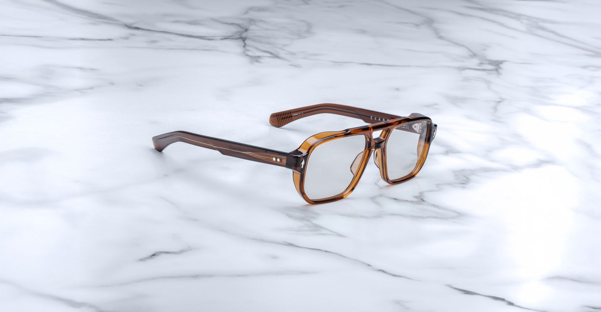 The Jacques Marie Mage Evans, a pair of oversized brown square acetate eyeglasses by Jacques Marie Mage, is displayed on a white marble surface.