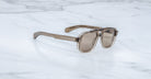 The Jacques Marie Mage Evans sunglasses feature translucent brown rectangular lenses and an oversized acetate frame, resting on a white marble surface and evoking the brand’s signature handcrafted optical style.