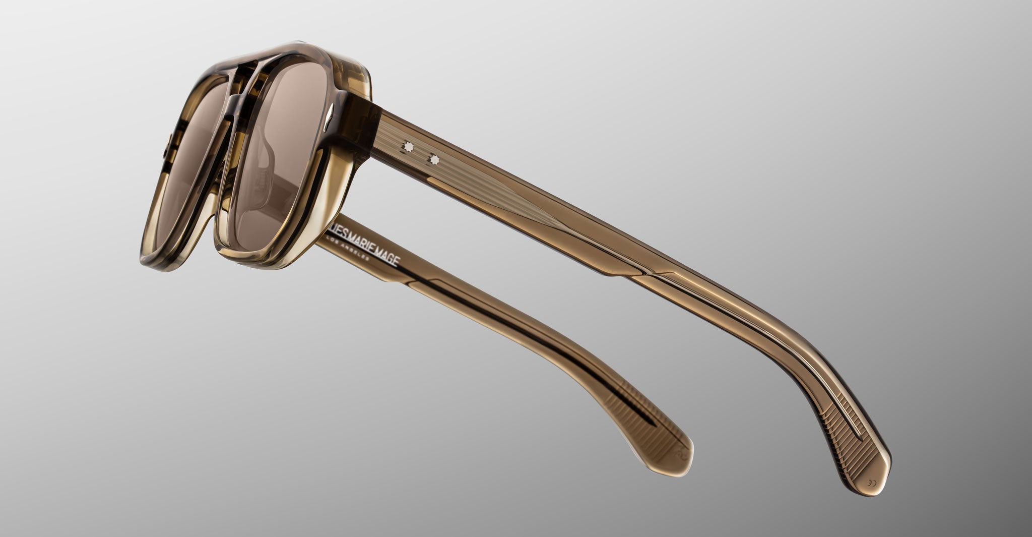 Close-up of Jacques Marie Mage Evans sunglasses, featuring an oversized brown translucent acetate frame and tinted lenses, displayed from a side angle against a gradient gray background, echoing the handcrafted quality of Jacques Marie Mage eyewear.