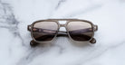 The Jacques Marie Mage Evans sunglasses by Jacques Marie Mage feature an oversized brown acetate square frame with dark lenses, displayed on a white marble surface.