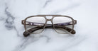 The Jacques Marie Mage Evans eyeglasses by Jacques Marie Mage feature an oversized gray acetate frame with thick rims and rectangular lenses, shown resting on a white marble surface.